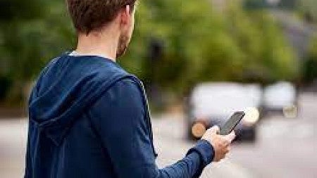 A man holding a cell phone in his right hand stands on a sidewalk and awaits an approaching car. 