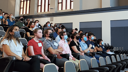 A group of masked students sit in a classroom listening intently to an unseen speaker. 