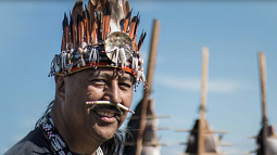 Smiling with the sun illuminating the left of his face, Jason Younker stands against a blue sky wearing Coquille regalia. 