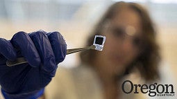 A gloved hand holding tweezers that grasp a tiny chip is in focus against a blurred background 