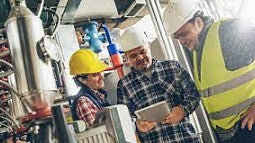 Three professionals wear hardhats as they talk animatedly.