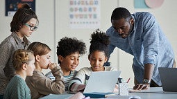 Young students congregate around a computer, smiling, as their teacher provides support. 