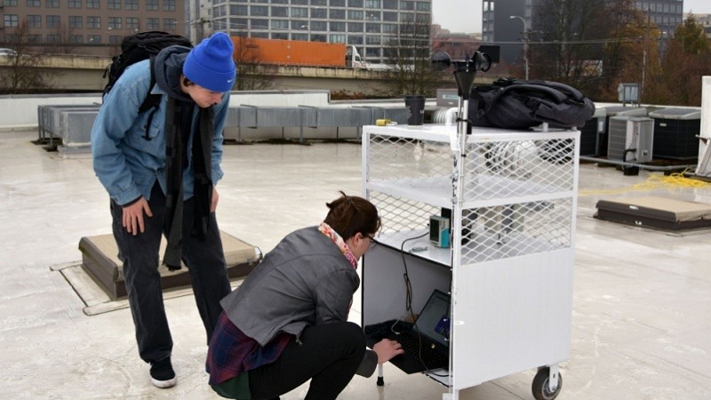 Elise Braun, right, and Thomas Cooper collect data from a weather station on the roof of Opsis Architecture, as a semitruck drives by on Interstate 405.