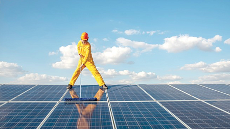 A person wearing a full-body yellow work suit and orange hard hat cleans solar panels using a special mop