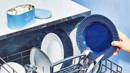A hand holding a blue dinner plate hovers above a dishwashing machine full of white and blue plates. 