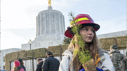 A young person holding a tree seedling and wearing a red hard hat looks to the left and smiles slightly. In the background, several others are congregating near a truck loaded with hay. Further back sits the Oregon State Capitol building.  