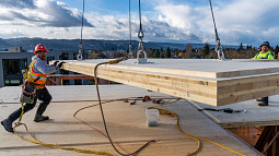 Two construction workers help build a roof, guiding to the appropriate place a stack of wood that is suspended by a system of cables. Other buildings, trees, and blue sky fill the background. 