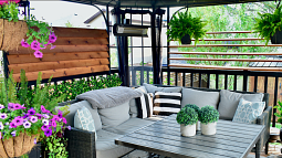 A sun patio with passive cooling devices is shown. The patio holds a plush couch with several throw pillows, planters with flowers and greenery, and a coffee table adorned with small shrubs. 