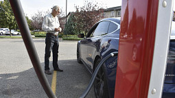 A person stands facing their electric car as it charges in a parking lot. 