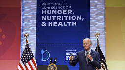 Joe Biden delivers remarks at the White House conference on hunger, nutrition, and health in Washington, DC. In front of Joe is a podium with a U.S. seal and two microphones, though Joe also holds a microphone in his left hand. Behind Joe are two national flags and a banner displaying the conference title and logo.  