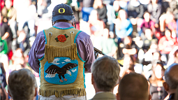 A person wearing an artistic buckskin vest and a backwards UO hat stands and faces a blurred crowd