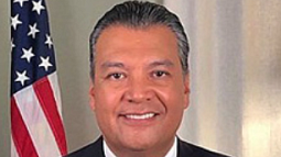 U.S. Senator Alex Padilla directly faces the camera and smiles. An American flag sits in the background over Padilla's right shoulder.