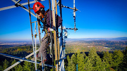 Silas Thoms, Multi-Hazards Field Technician with the Oregon Hazards Lab, is perched high up in a telecommunications tower overlooking an wide expanse of forestland.