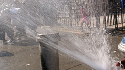 A person uses their hand to direct water from a fire hydrant toward a group of people standing on a sidewalk trying to cool down in the harsh heat