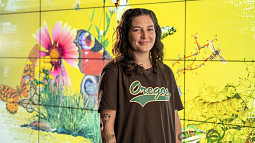 Jordan Rodriguez wears a University of Oregon shirt and poses before a digital display representing biodiversity through depictions of various wildlife
