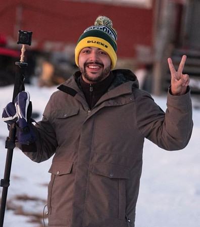 Researcher Danny Pimentel in a snowy outdoor location carrying a tripod and wearing a UO hat.